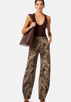 Donna con top marrone senza maniche, pantaloni ampi con fantasia, che tiene una grande borsa tote in pelle marrone su una spalla.