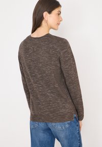 Ruskea teksturoitu neulepaita, jossa on pyöreä pääntie ja pitkät hihat, hienovaraisella kuviolla ja sivuhalkioilla, päällä siniset farkut.