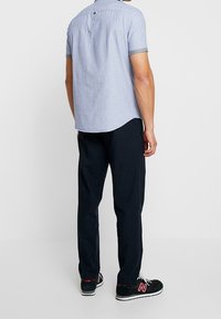Camicia azzurra a maniche corte con colletto, abbinata a pantaloni scuri e sneakers nere con dettagli rossi, vista da dietro.