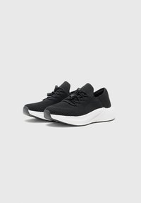 Chaussures de sport noires avec une tige en tricot texturé et une semelle blanche rembourrée. Elles présentent un bout rond, un design à lacets et une languette au talon arrière.