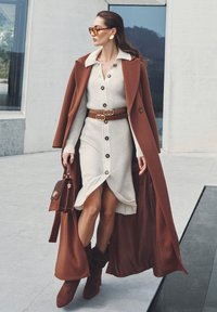 Bruine overjas, crèmekleurige geribde jurk met knopen, bruine riem, bijpassende laarzen en handtas. Accessoires omvatten oversized zonnebril en oorbellen.