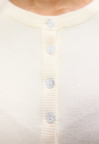 Cardigã de cor creme com um decote canelado, apresentando cinco botões redondos e claros na frente. Textura suave, material de malha leve.