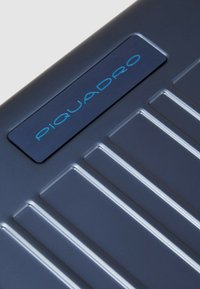 Valigia rigida blu scuro con scanalature orizzontali, una piastra logo metallica e una superficie liscia.