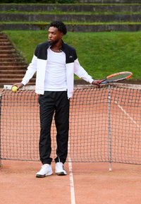 Jucător de tenis îmbrăcat într-o jachetă cu fermoar alb-negru, cu tricou alb și pantaloni negri, ținând o minge de tenis și o rachetă lângă un fileu.