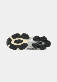 Semelle de chaussure de sport, avec une semelle extérieure en caoutchouc noir texturé présentant diverses formes, reliée à une semelle intermédiaire blanche avec des trous de ventilation.