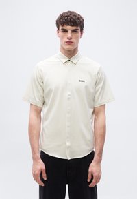 Νέος άνδρας που φοράει κοντομάνικο, ανοιχτό μπεζ πουκάμισο με ρίγες και γιακά, με το λογότυπο "BOSS" στο στήθος και μαύρο παντελόνι, στέκεται μπροστά σε λευκό φόντο.