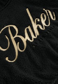 Tessuto in pile nero con una superficie testurizzata, caratterizzato da un prominente testo ricamato in oro che recita "Baker" in un font corsivo.