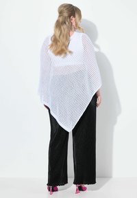 Biały dziergany poncho z wzorem w diamenty swobodnie opada na czarne spodnie o szerokich nogawkach. Różowe szpilki widoczne na dole. Miękka faktura.