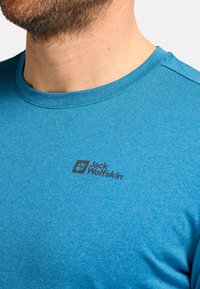 Uomo che indossa una t-shirt blu a girocollo con un logo nero di Jack Wolfskin sul petto, visibile dalla nuca fino a metà torso.