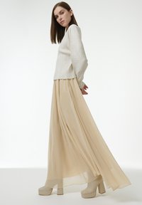 Frau mit glattem braunem Haar, die ein cremefarbenes geripptes Langarmoberteil, einen fließenden beigen Maxirock und beigen Plateauschuhen trägt, stehend im Profil.
