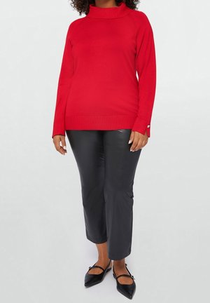 Rød strikket sweater med høj krave og ribbet kant, parret med sorte bukser i kunstlæder, der slutter kort. Skoene er spidse sorte flade sko.