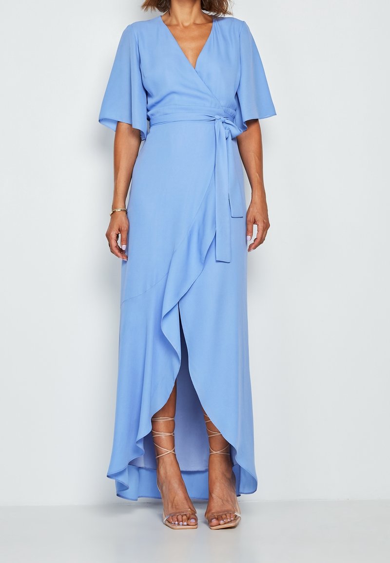 Femme portant une robe portefeuille bleu clair avec manches volantées et ourlet asymétrique à volants, associée à des sandales à talons hauts nude à lanières.