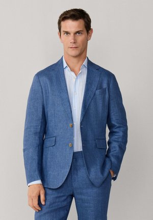 Homme portant un costume en lin bleu à deux boutons, associé à une chemise habillée à rayures bleu clair, debout avec une main dans la poche.