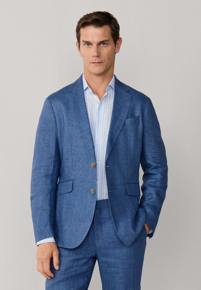 Mann trägt einen blauen Leinenanzug mit zwei Knöpfen, kombiniert mit einem hellblauen gestreiften Hemd, steht mit einer Hand in der Tasche.