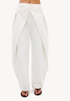 Pantalon blanc à jambes larges avec panneaux avant qui se chevauchent et plis latéraux, porté avec des tongs foncées sur un fond neutre.