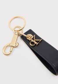 Χρυσό μπρελόκ με κούμπωμα προσαρτημένο σε μαύρο δερμάτινο λουράκι με χρυσά γούρια λογότυπου CK σε λευκό φόντο.