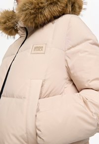Chaqueta acolchada beige que presenta una capucha forrada de piel desmontable, un parche de marca y una textura acolchada con cierre frontal y bolsillos laterales.