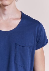 Tyrkysové tričko s krátkým rukávem vyrobené z měkké tkaniny, s náprsní kapsou a kulatým výstřihem. Hladká textura a ohrnuté rukávy.