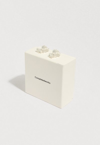 Brincos de flor transparentes em forma de tacha posicionados em cima de uma caixa branca rotulada "Completedworks", apresentando um design minimalista e textura suave.