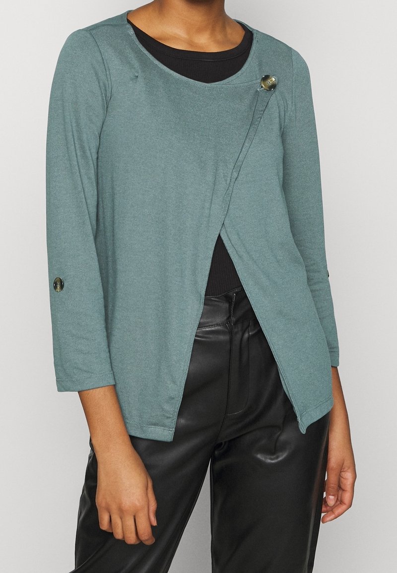 Femme portant un haut à manches longues style cache-cœur bleu sarcelle avec des boutons sur une chemise noire et un pantalon en cuir noir.