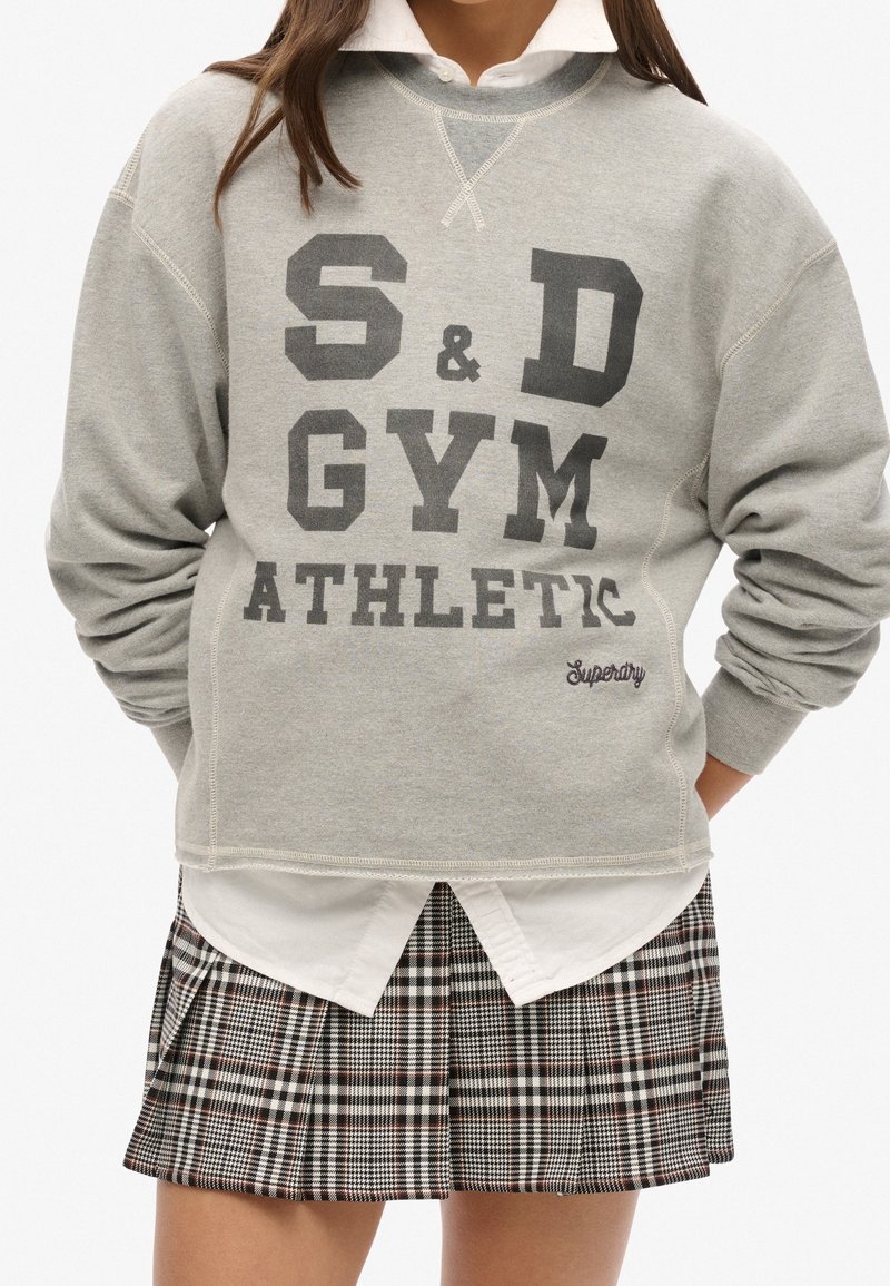 Sweat-shirt gris avec le texte noir "S&D GYM ATHLETIC". Superposé sur une chemise blanche à col, accompagné d'une jupe plissée à carreaux.