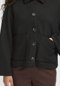 Veste noire à côtes, avec de grands boutons et deux poches plaquées à l'avant, portée par une personne avec une main dans une poche.