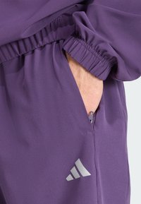 Lila Sportjacke mit elastischen Bündchen, einer seitlichen Tasche mit Reißverschluss und einem silbernen Logodetail im unteren Bereich. Glatte Textur.