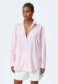 Light pink button-up shirt featuring white embroidered heart patterns, long sleeves, a pointed collar, and a relaxed fit. Casual design.