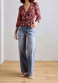 Květinový peplum top v červené, růžové a černé barvě, s výstřihem do V a tříčtvrtečními rukávy, doplněný světle modrými širokými džíny a bílými sandály na platformě.