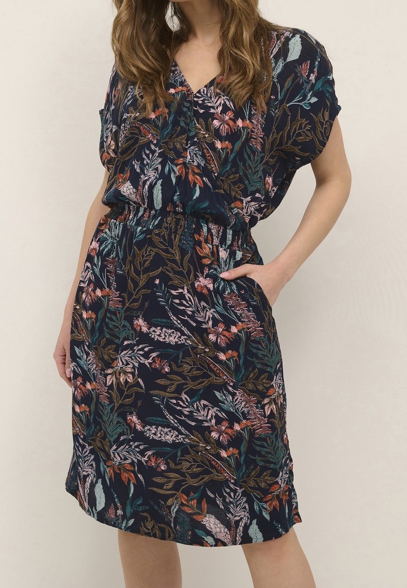 Femme portant une robe bleu marine à manches courtes et imprimé floral multicolore, main reposant dans la poche sur fond beige.
