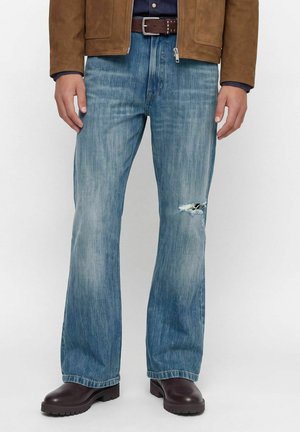 Homme portant un jean bleu délavé avec un trou au genou droit, ceinture marron foncé, veste marron et bottes marron foncé épaisses.