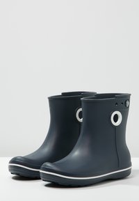 Bottes de cheville en caoutchouc noir avec des accents blancs et des trous circulaires sur les côtés. Texture lisse avec un bout arrondi et une bande de semelle blanche.