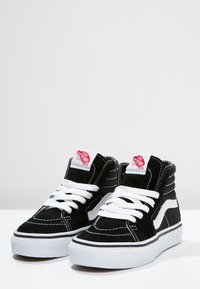 Czarne zamszowe sneakersy typu high-top z białymi sznurowadłami i gumowymi podeszwami. Zawierają czerwony znacznik logo i białą falistą wzór po boku.