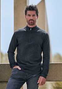 Dunkelgrauer Reißverschluss-Pullover mit hohem Kragen, langen Ärmeln und schlanker Passform, kombiniert mit dunklen Jeans. Glatte Textur, minimalistisches Design.