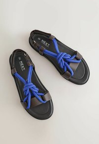 Sandales noires avec des lanières bleues nouées et des accents marron, indiquées en taille 6,5/40 avec la mention "NEXT Forever Comfort" sur la semelle intérieure.