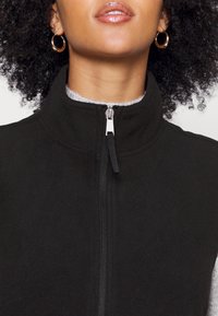 Svart fleeceväst med dragkedja, hög krage och silverfärgad dragkedja. Mjuk textur med kontrasterande innerv färg. Inga synliga fickor.