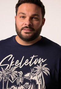 Camiseta de color azul marino hecha de algodón, que presenta un gran gráfico blanco de un esqueleto con palmeras y olas, impreso en la parte frontal.