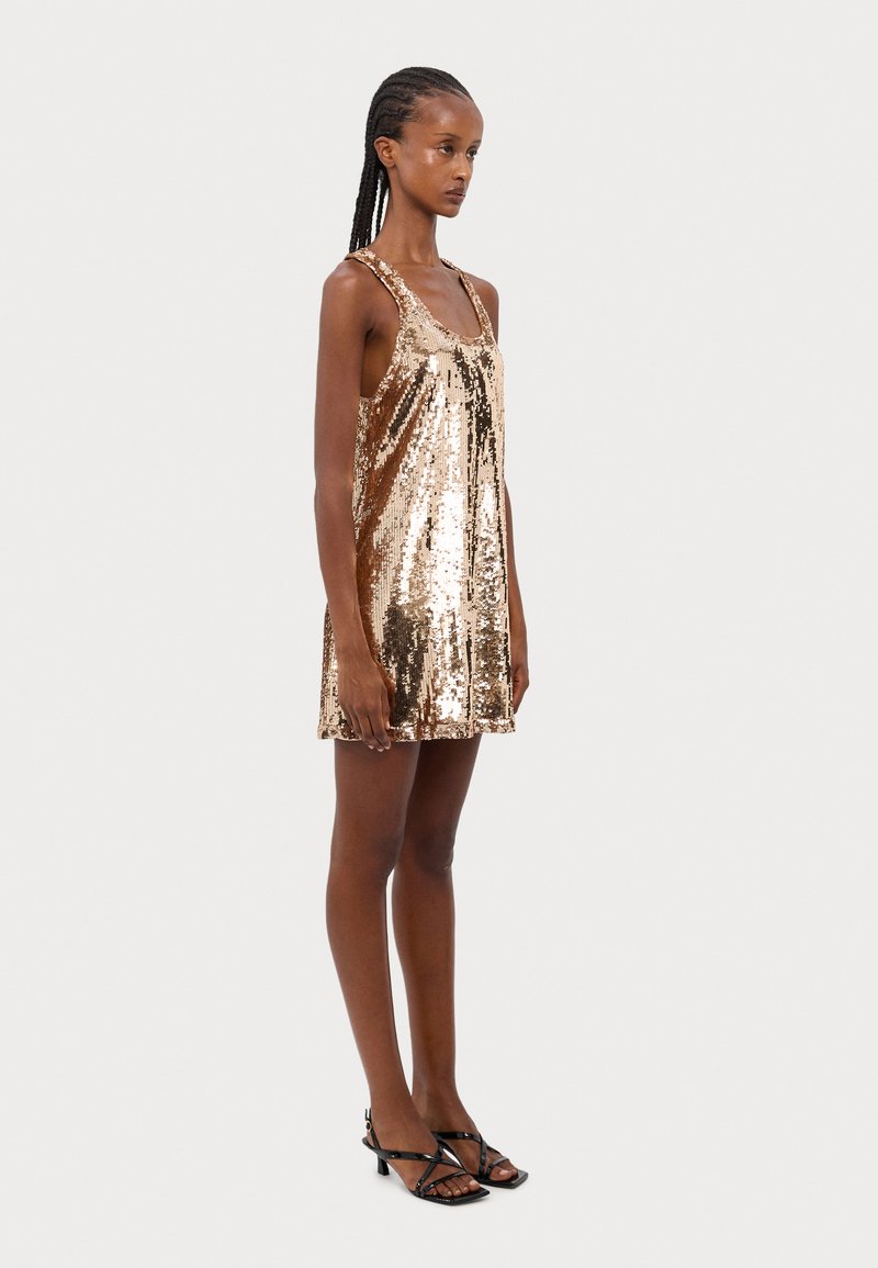 Robe en sequins dorés avec une coupe ample, présentant un col rond et des bretelles, associée à des talons noirs à lanières.