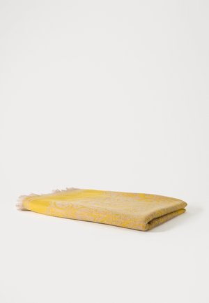 Tissu plié jaune et beige à motifs avec bords frangés, posé à plat sur une surface blanche.