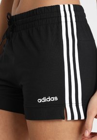 Pantalones cortos deportivos negros con cinturilla elástica, que cuentan con tres rayas verticales blancas en el costado y un logo de Adidas en blanco en el dobladillo.