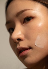 Twarz nakładająca na skórę białą krem, posiadającą gładką, lekko błyszczącą teksturę. Naturalny makijaż podkreśla rysy twarzy w miękkim świetle.