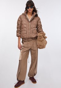 Giacca imbottita marrone con design trapuntato, tasche laterali e cerniera. Pantaloni color beige in raso con coulisse. Il modello tiene in mano un peluche di coniglio soffice.