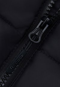 Zbliżenie na czarną, pikowaną tkaninę kurtki z czarnym zamkiem błyskawicznym; zawiera teksturowany uchwyt zamka oraz oznaczoną metkę z logo „Reebok”.