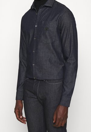 Homme portant une chemise en jean sombre à boutons avec poche poitrine, associée à un jean en jean sombre assorti, sur un fond uni.