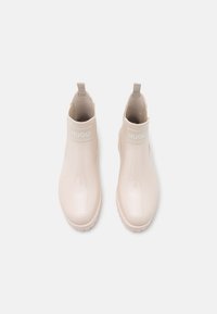 Bottines en caoutchouc beiges avec une finition brillante, dotées d'une bande logo en haut, de languettes de tirage et d'une semelle à motif texturé.