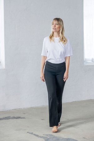 Mujer rubia descalza caminando en interiores sobre un suelo de hormigón, vistiendo una camisa blanca y pantalones negros, mirando ligeramente hacia arriba a la derecha.