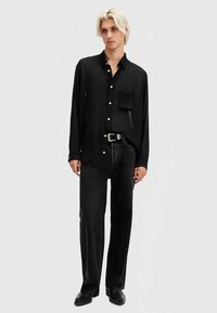 Melnas garām piedurknēm krekls ar priekšējo kabatu un pogu aizdari, kombinēts ar tumšām džinsa biksēm un melniem apaviem, pelēka fona priekšā.