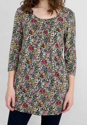 Frau trägt eine langärmelige Tunika mit dichtem floralen Muster in Weiß, Grün, Gelb und Rot auf dunklem Hintergrund.
