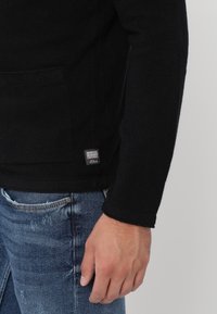 Musta neulepusero, jossa on teksturoitu pinta, etutasku, näkyvä tikkaus ja pieni merkkilappu helmassa. Yhdistetty sinisiin farkkuihin.