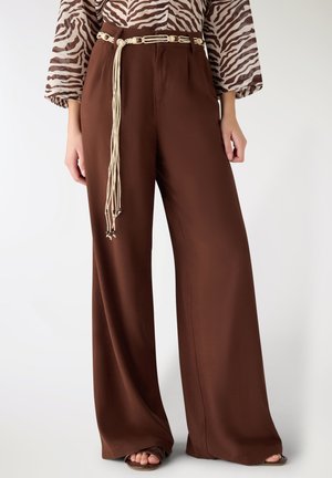 Donna che indossa pantaloni marroni a gamba larga con una cintura intrecciata color crema e una maglia a maniche lunghe con stampa zebra, in piedi contro uno sfondo chiaro e uniforme.